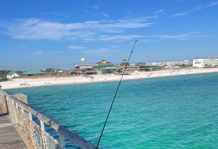 Destin real estate agent​view of Navarre Beach, Florida, from the pier, showing a fishing rod, turquoise Gulf water, and beachfront hotels and real estate.